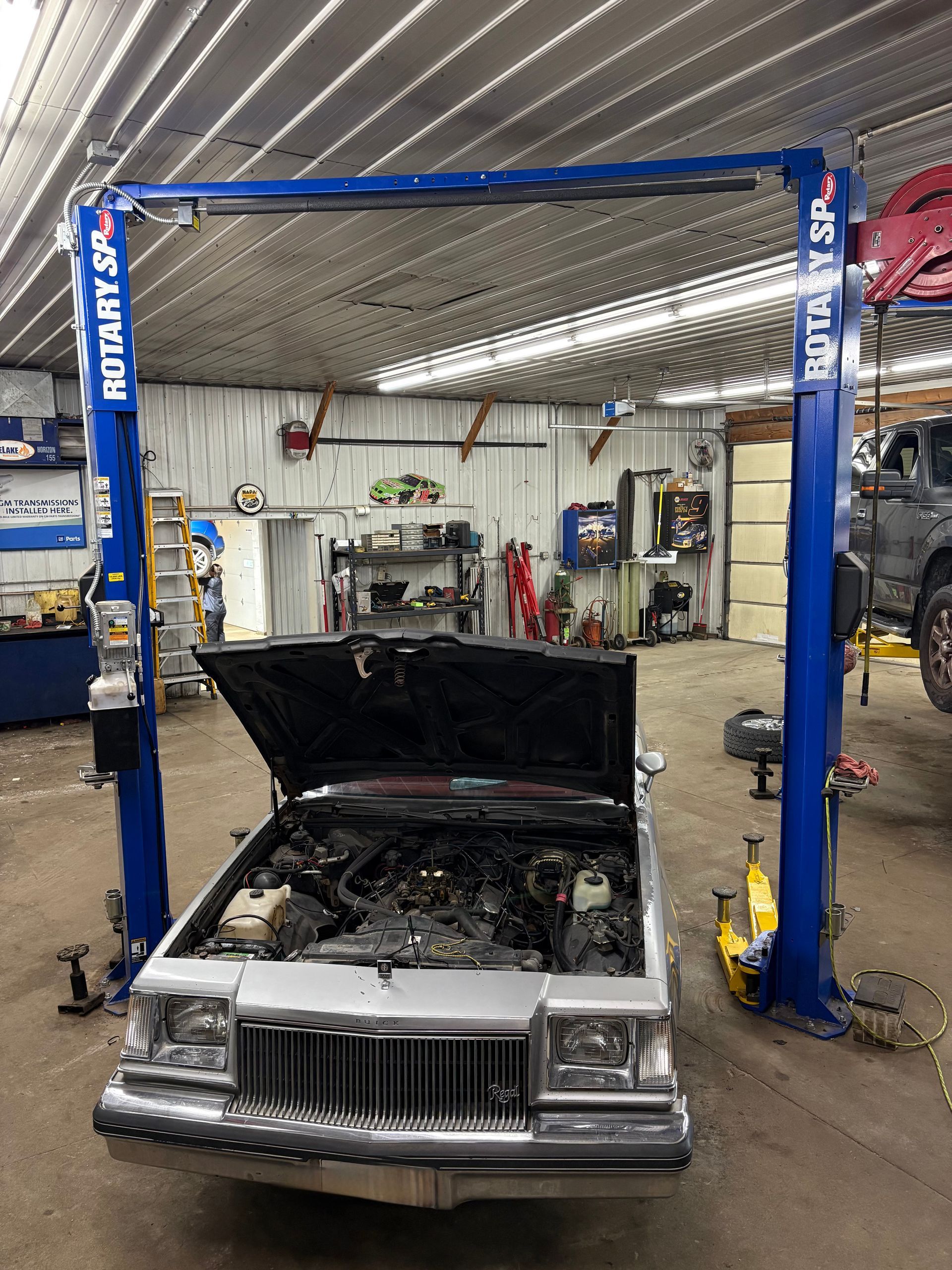 Old car with hood open under a blue auto repair lift in a garage.