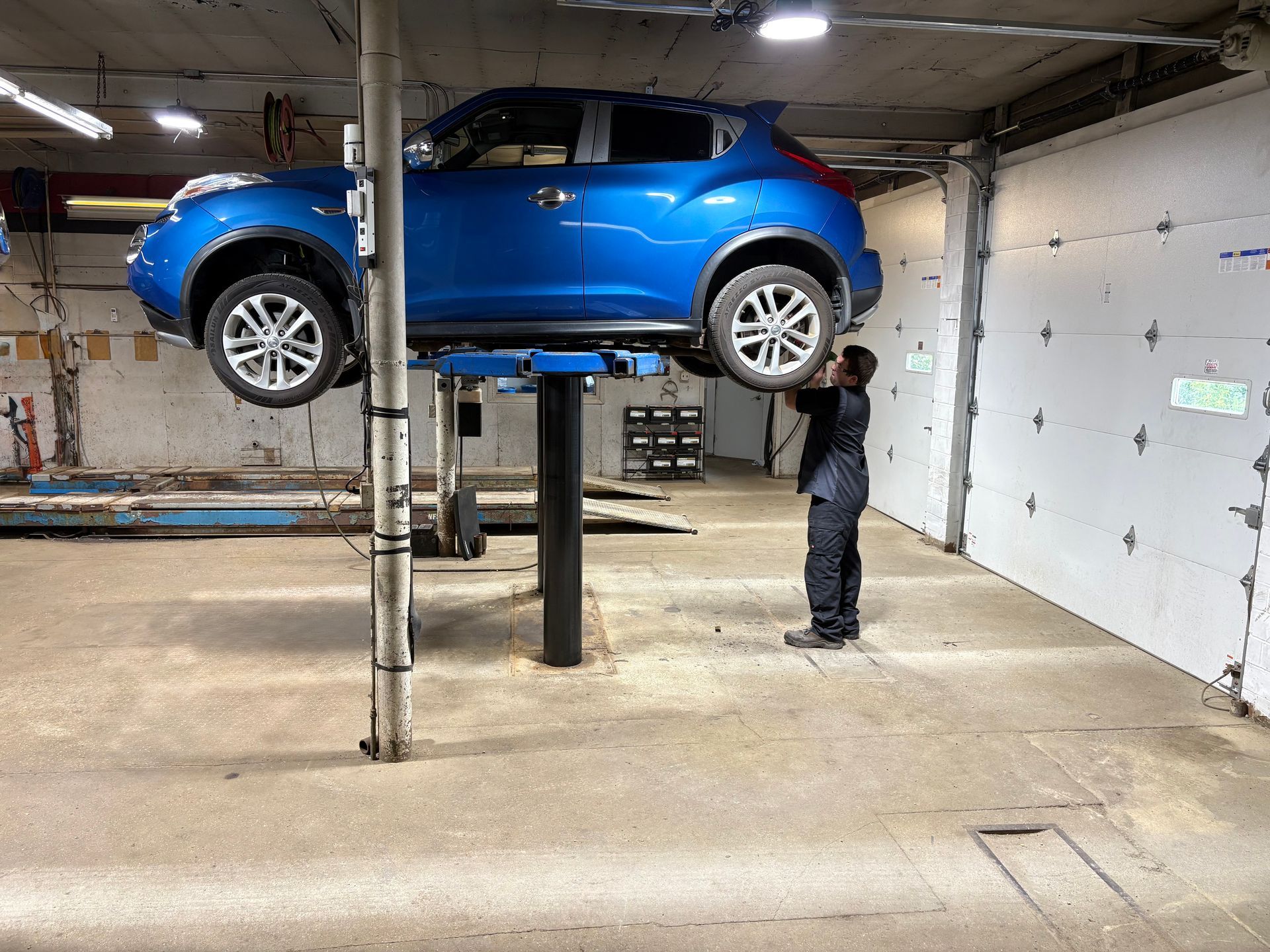 Blue car lifted on a garage hoist while a mechanic inspects it inside an auto shop.