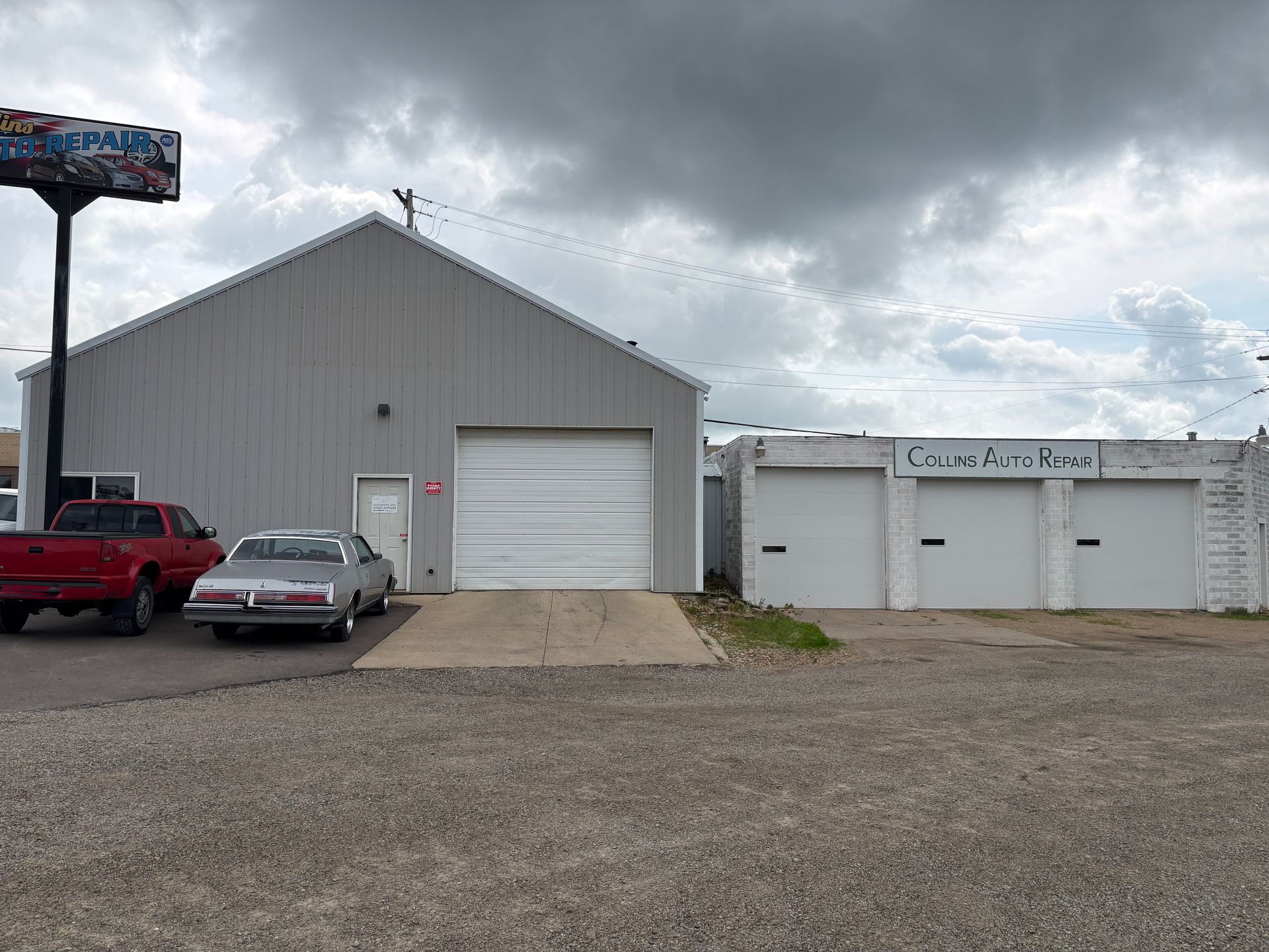 Gray metal building with garage door, parked red pickup and car, under cloudy sky