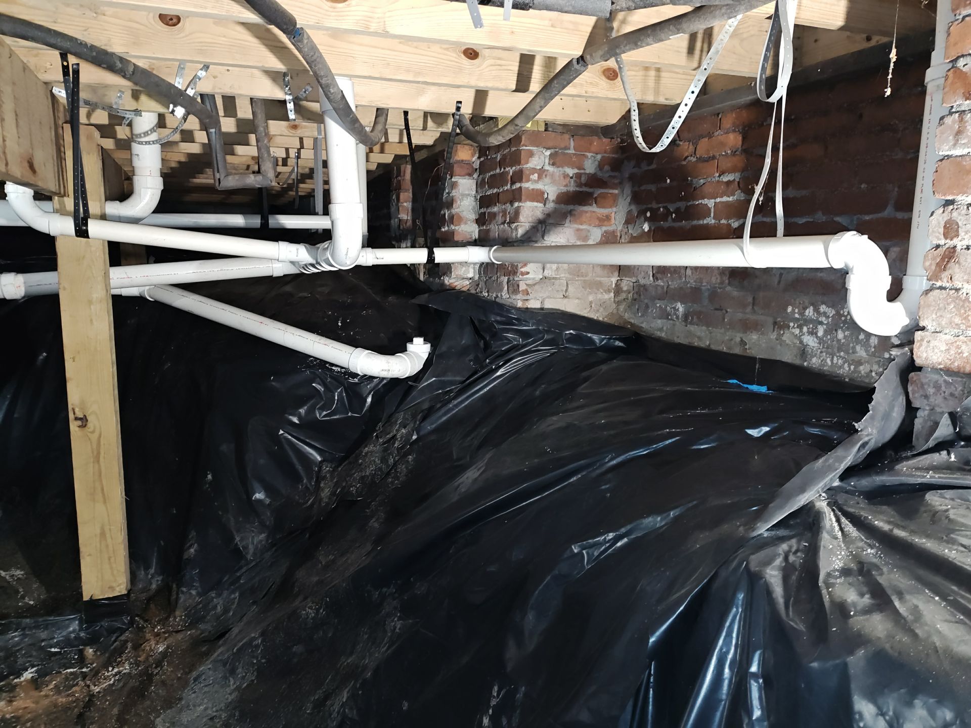 Crawlspace with exposed wooden joists, white plumbing pipes, and black plastic vapor barrier on the ground