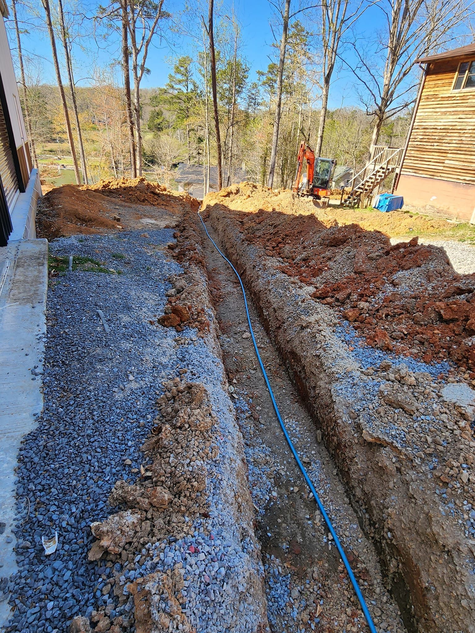Gravel trench beside a house with a blue pipe running through it and leaf-covered ground beyond