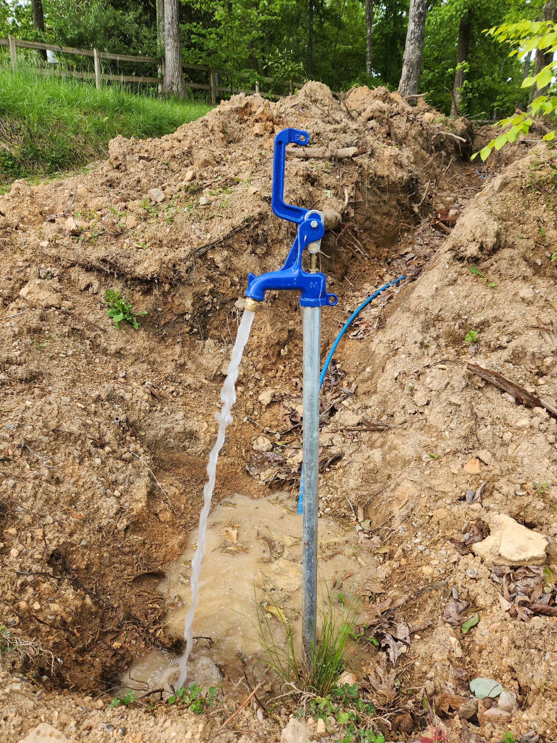 Blue water pump spraying water beside a muddy excavation site in a wooded area
