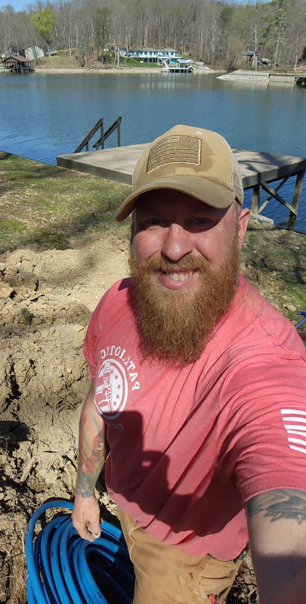 Selfie of a smiling bearded man in a red shirt and tan cap by a lake with a dock behind him