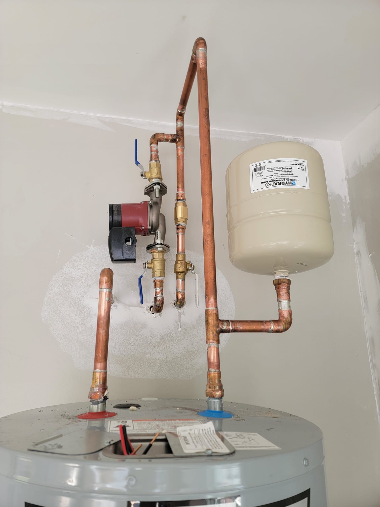 Copper pipes, a wall-mounted pump, and a beige expansion tank above a water heater in a utility room
