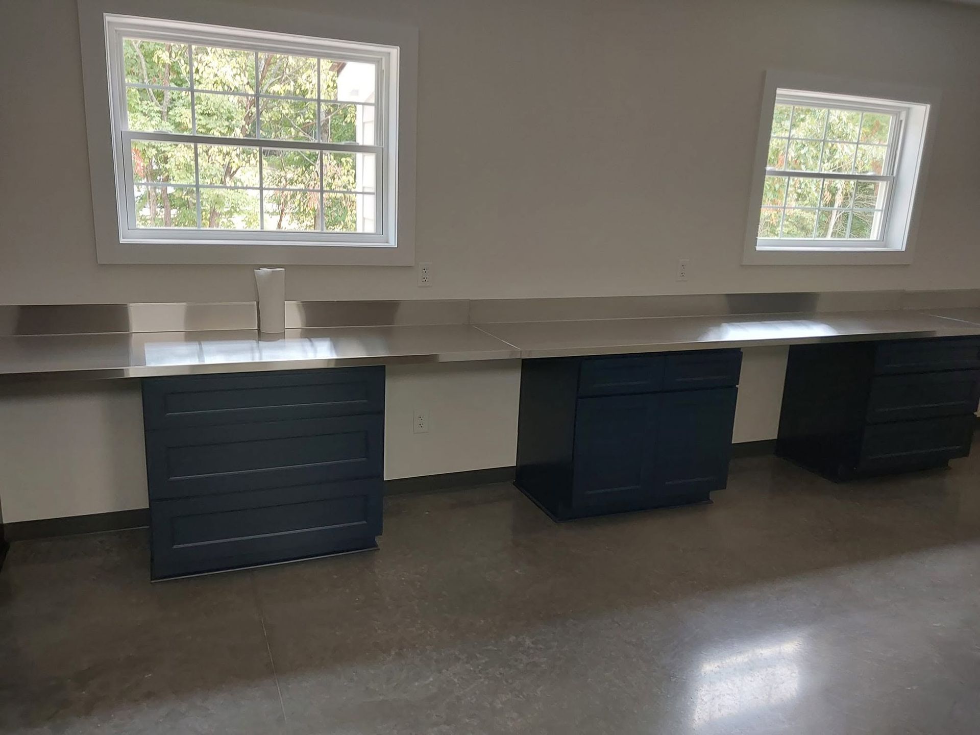 Empty room with two windows and three dark cabinets beneath a long white countertop