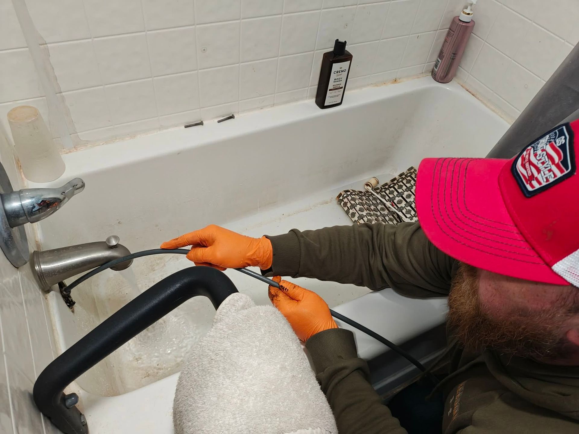 Person in orange gloves adjusting a black seat in a white bathtub, with bottles on the tub ledge.