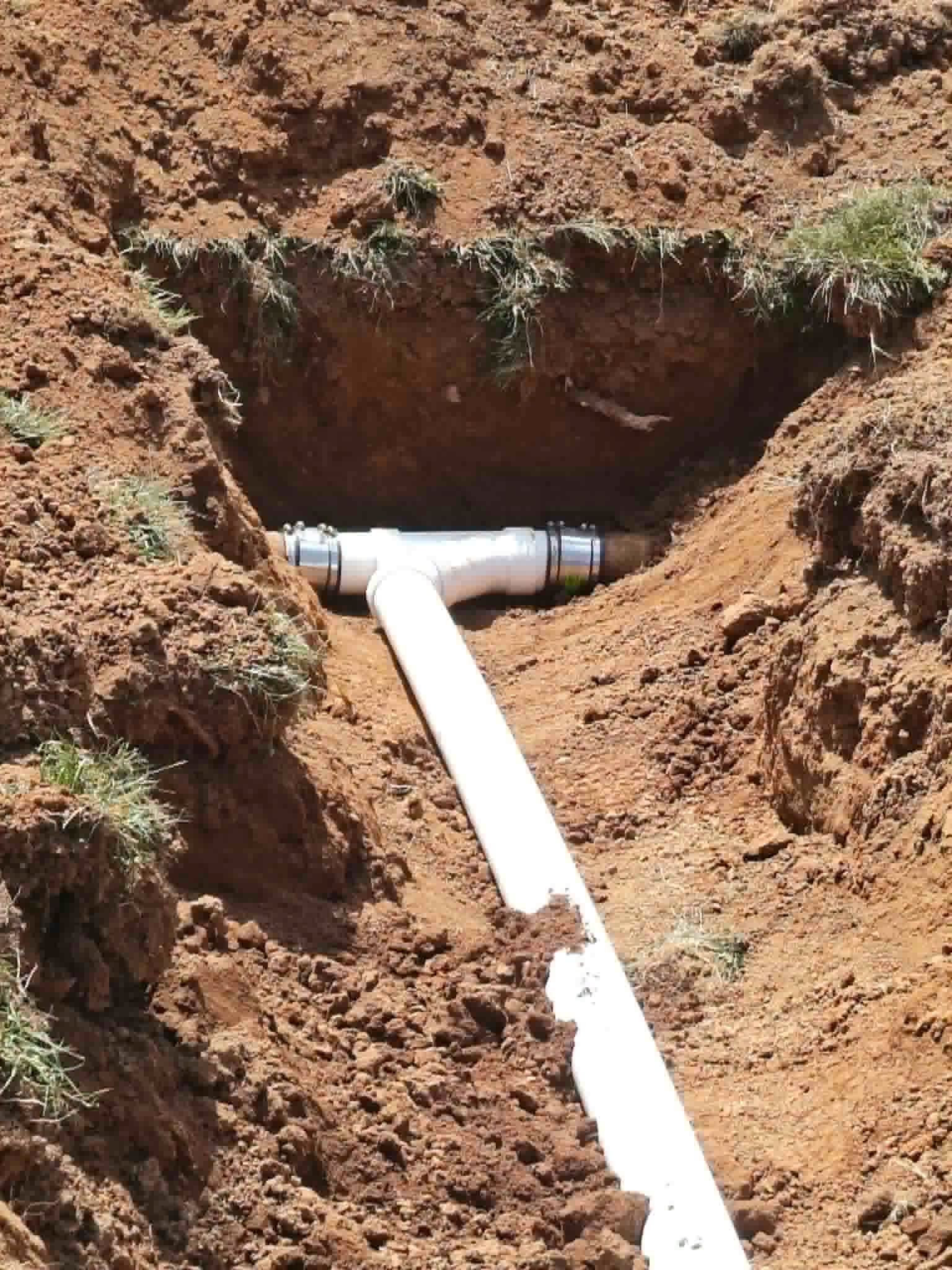 White pipe exposed in a muddy trench cut into reddish-brown soil, with a T-joint near the top