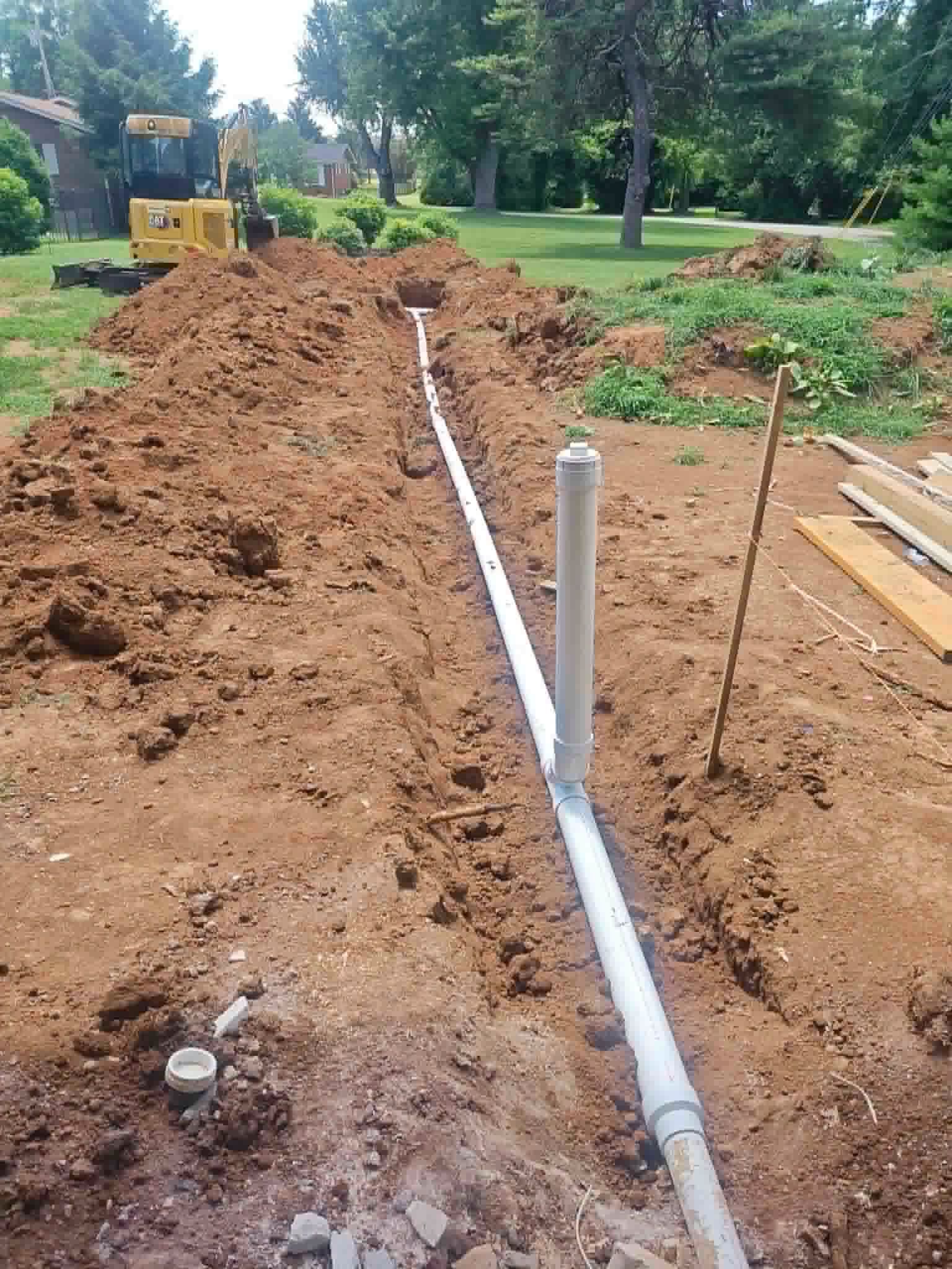 Trenched construction site with white PVC pipe, dirt piles, and an excavator in the background