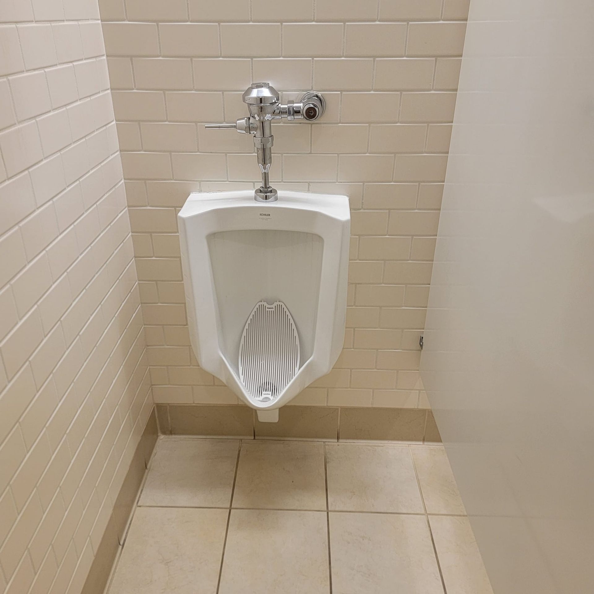 Urinal in a beige-tiled public restroom stall with chrome flush valve.