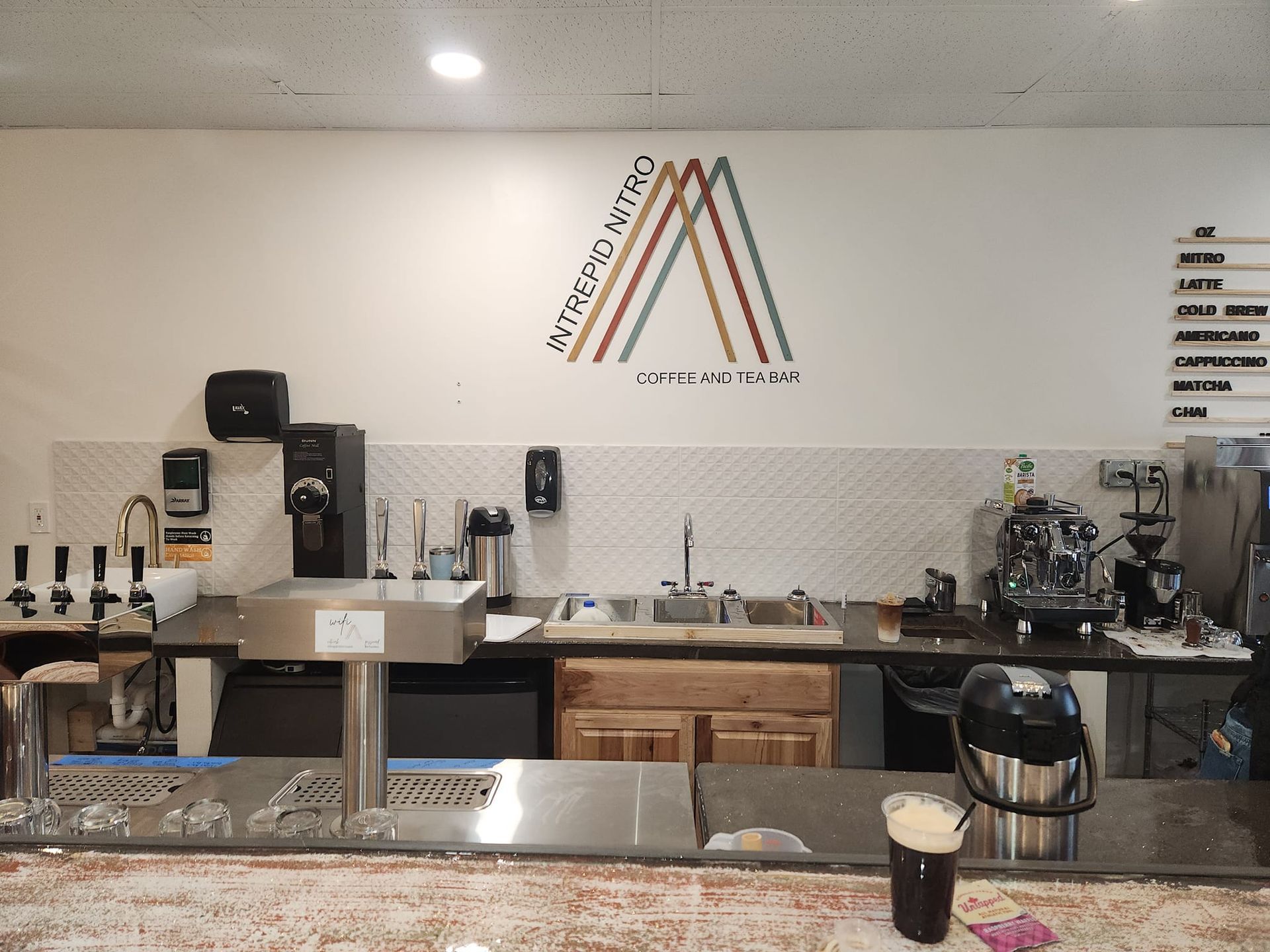 Coffee shop counter with espresso machines, cups, and a minimalist wall logo behind the bar.