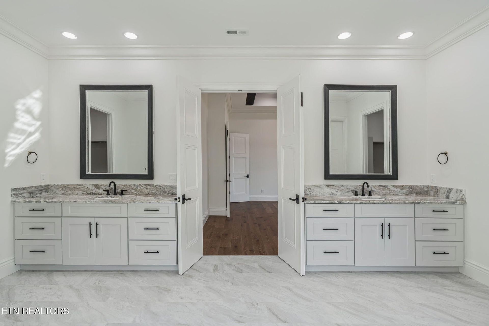 Bright bathroom with dual white vanities, large mirrors, marble countertops, and a doorway to a wood-floored hall.
