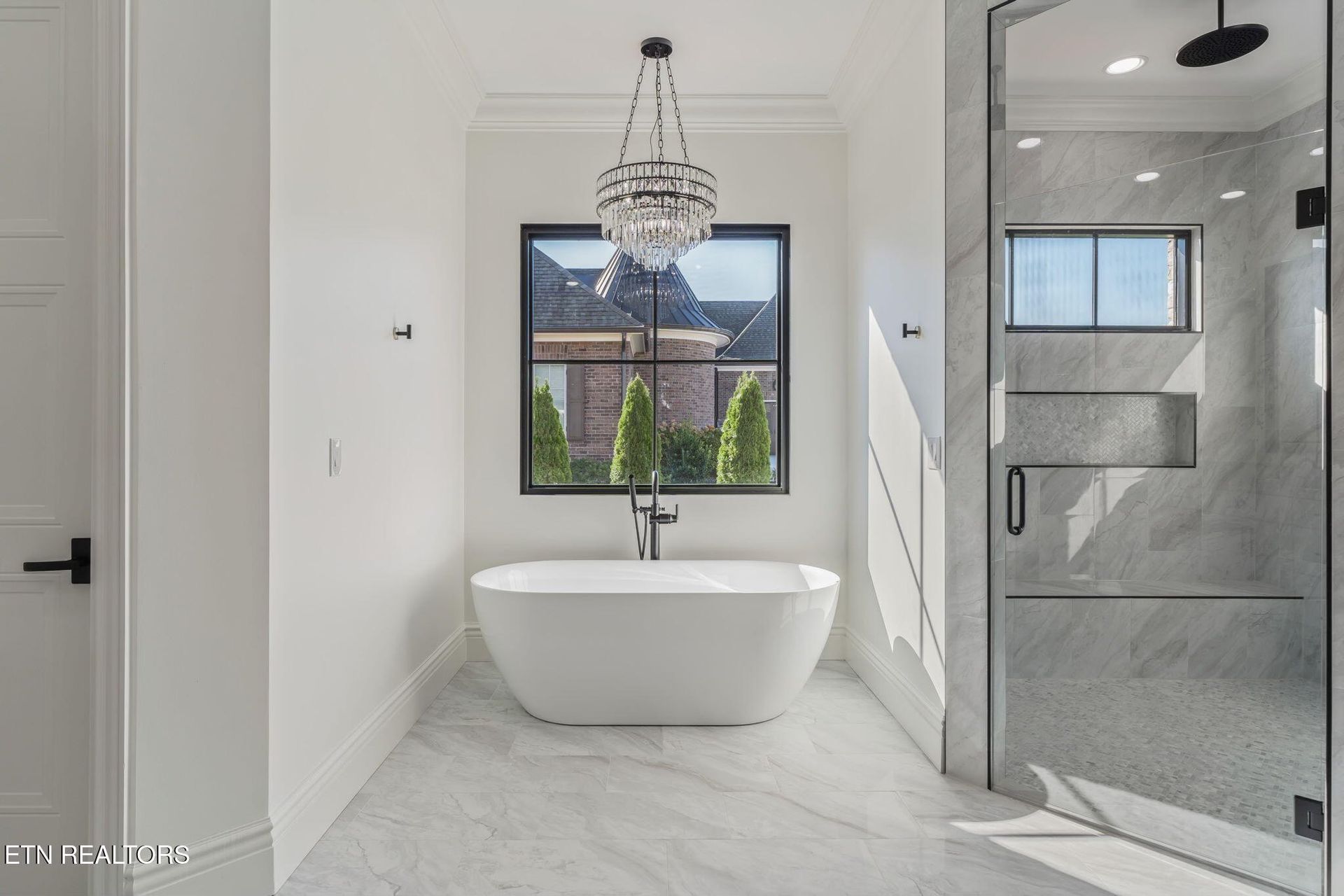 Bright modern bathroom with freestanding tub, chandelier, and glass shower.