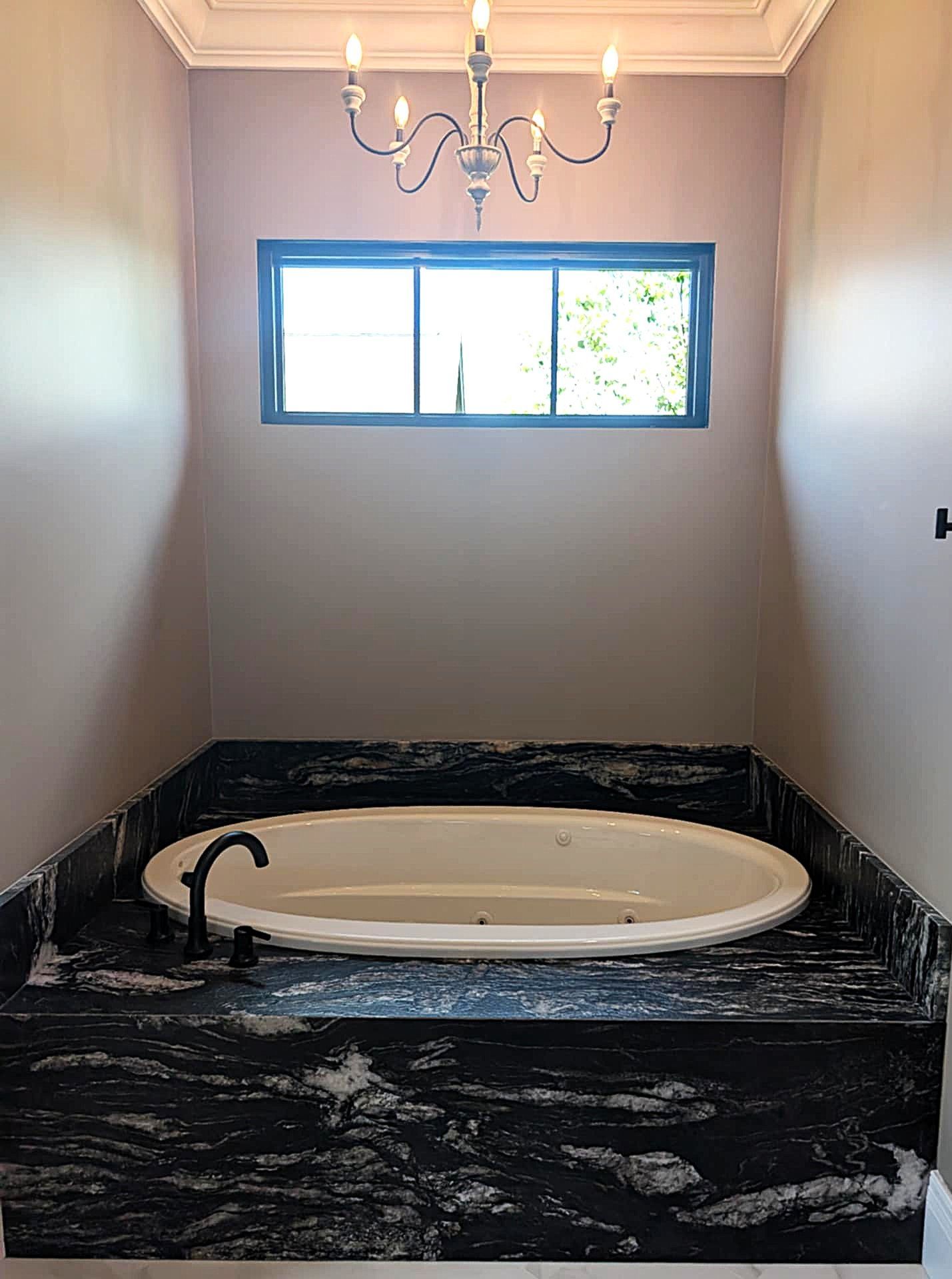 Bathroom with a black marble tub deck, oval bathtub, chandelier, and small window above.