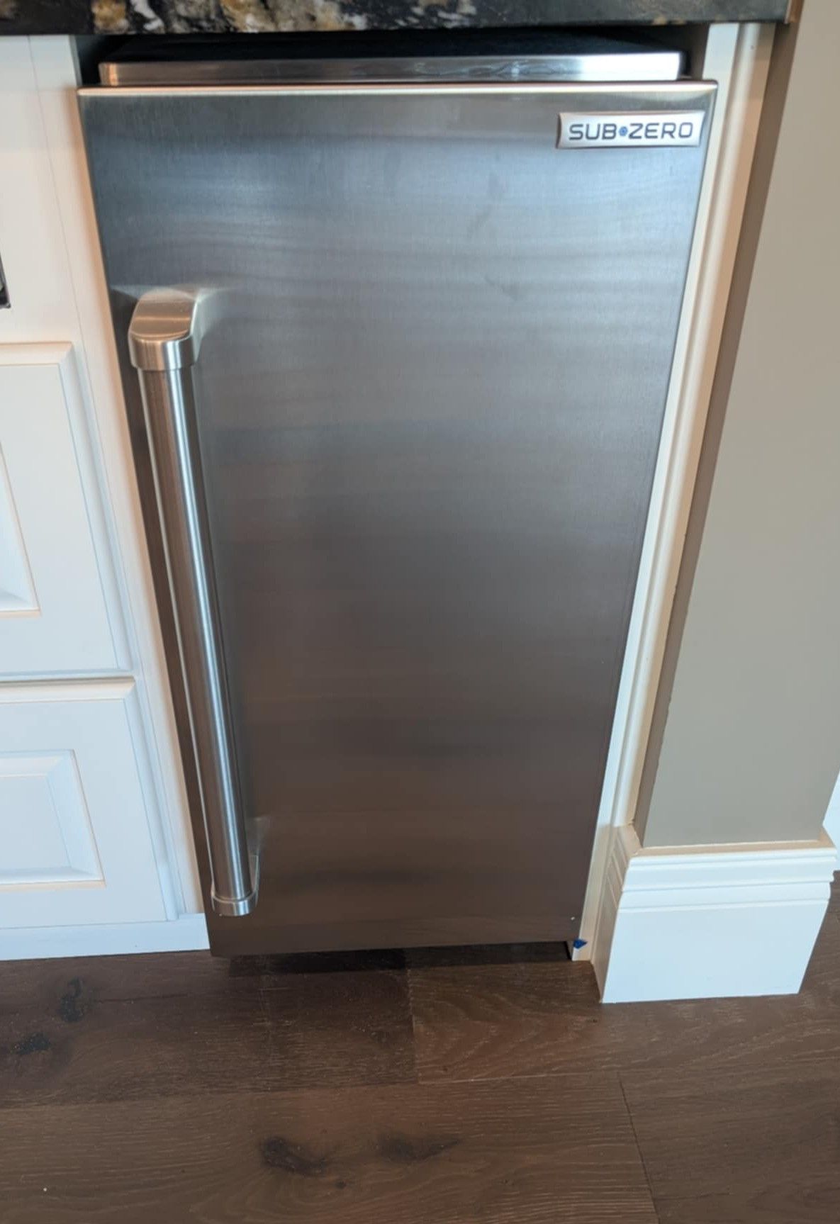 Stainless steel undercounter refrigerator with a long vertical handle, built into a kitchen cabinet opening.