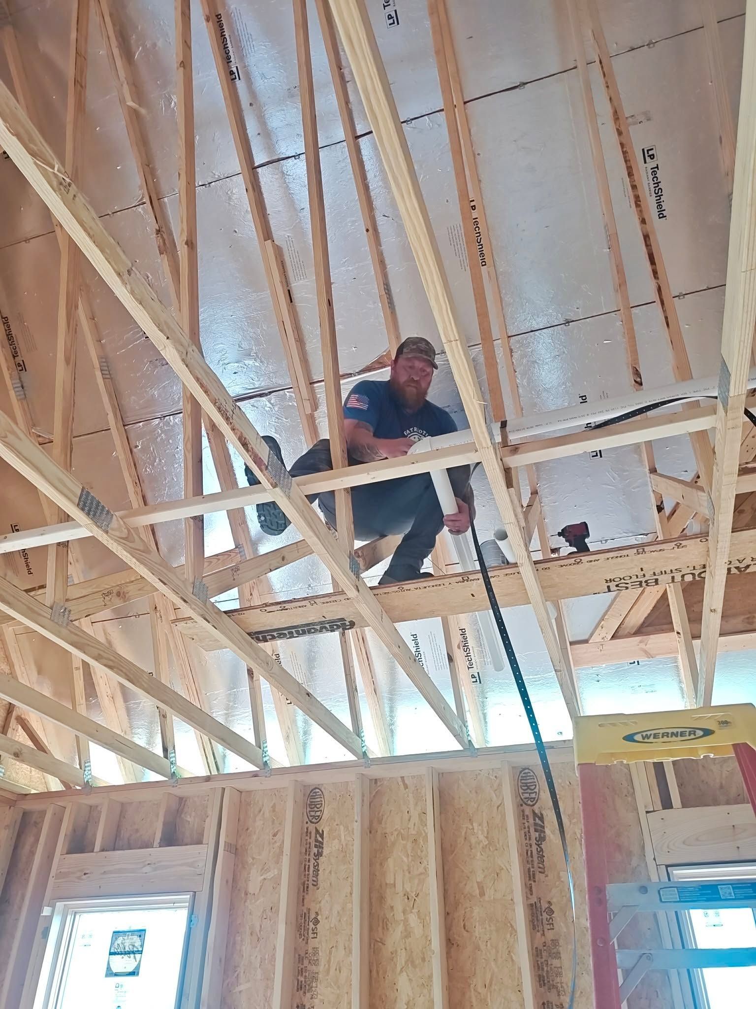 Worker on unfinished wood-framed ceiling, crouching among roof trusses at a construction site.