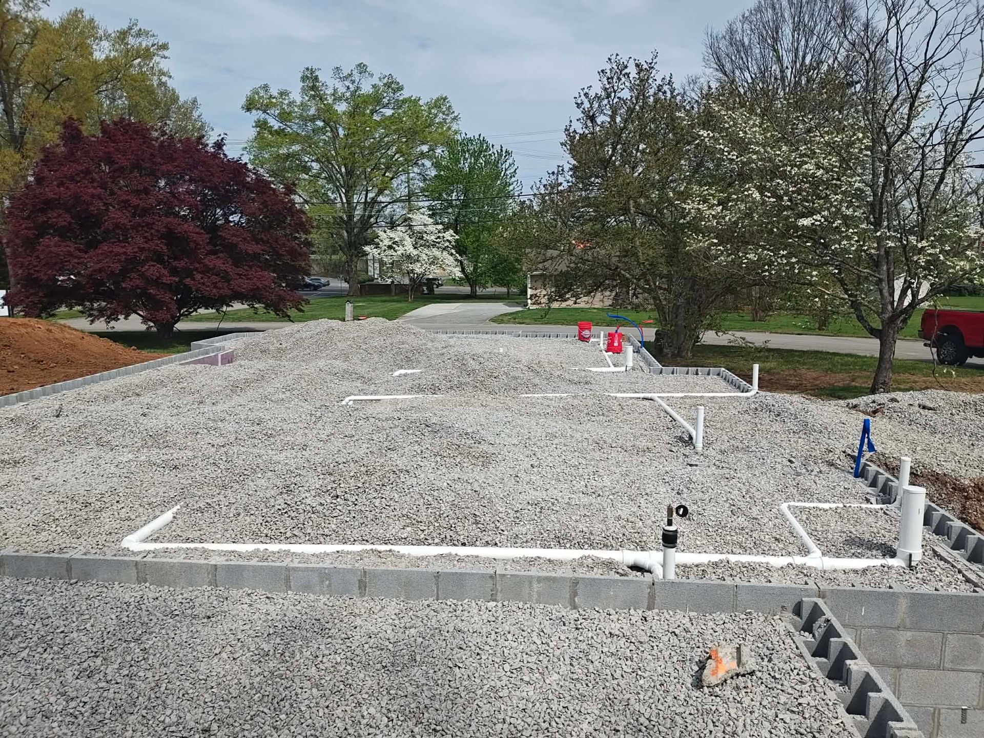 Gravel construction site with white foundation forms and red flags, bordered by trees and grass.