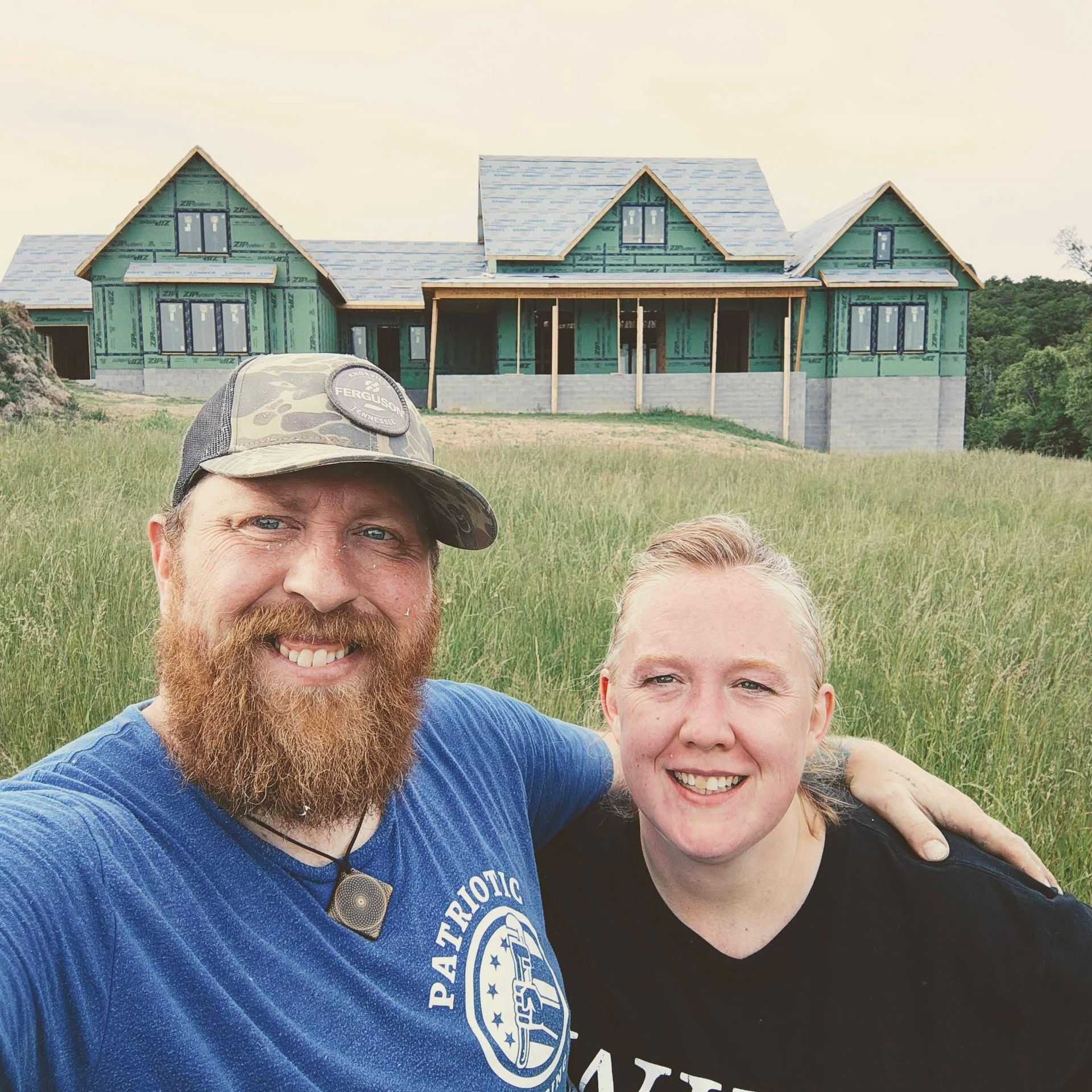 Couple smiling in front of a green cabin on a grassy hillside, with trees in the background.