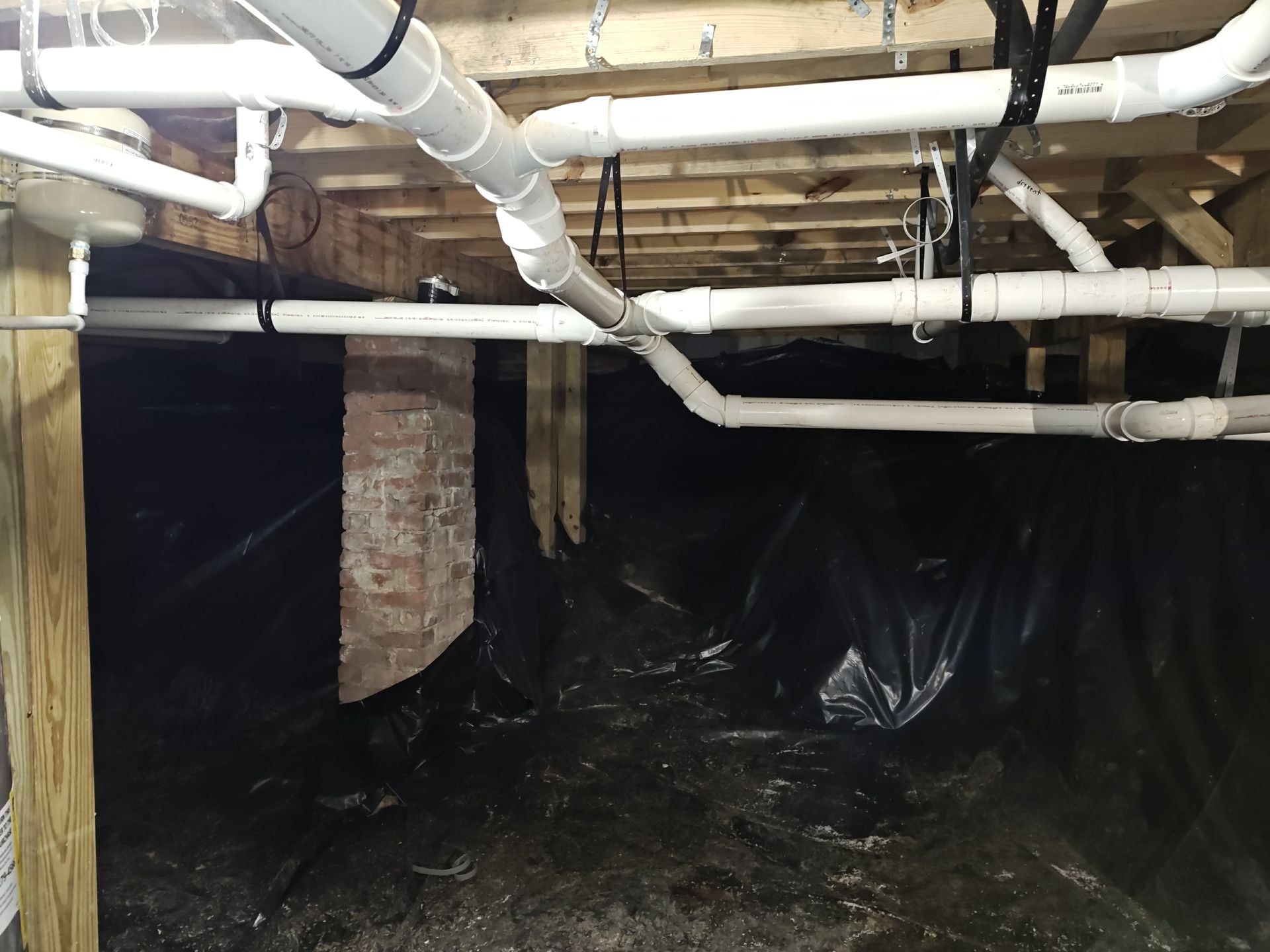 Crawl space with exposed pipes, black vapor barrier, and damp dirt floor under a house