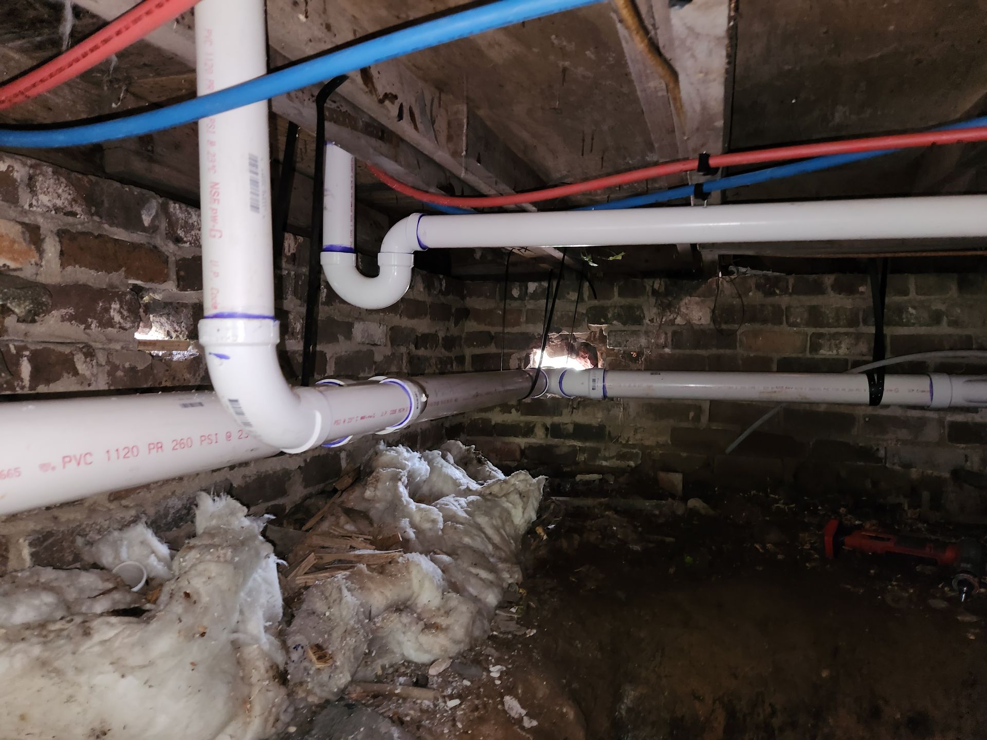 Damp crawl space with exposed white pipes, debris, and standing water under a brick foundation