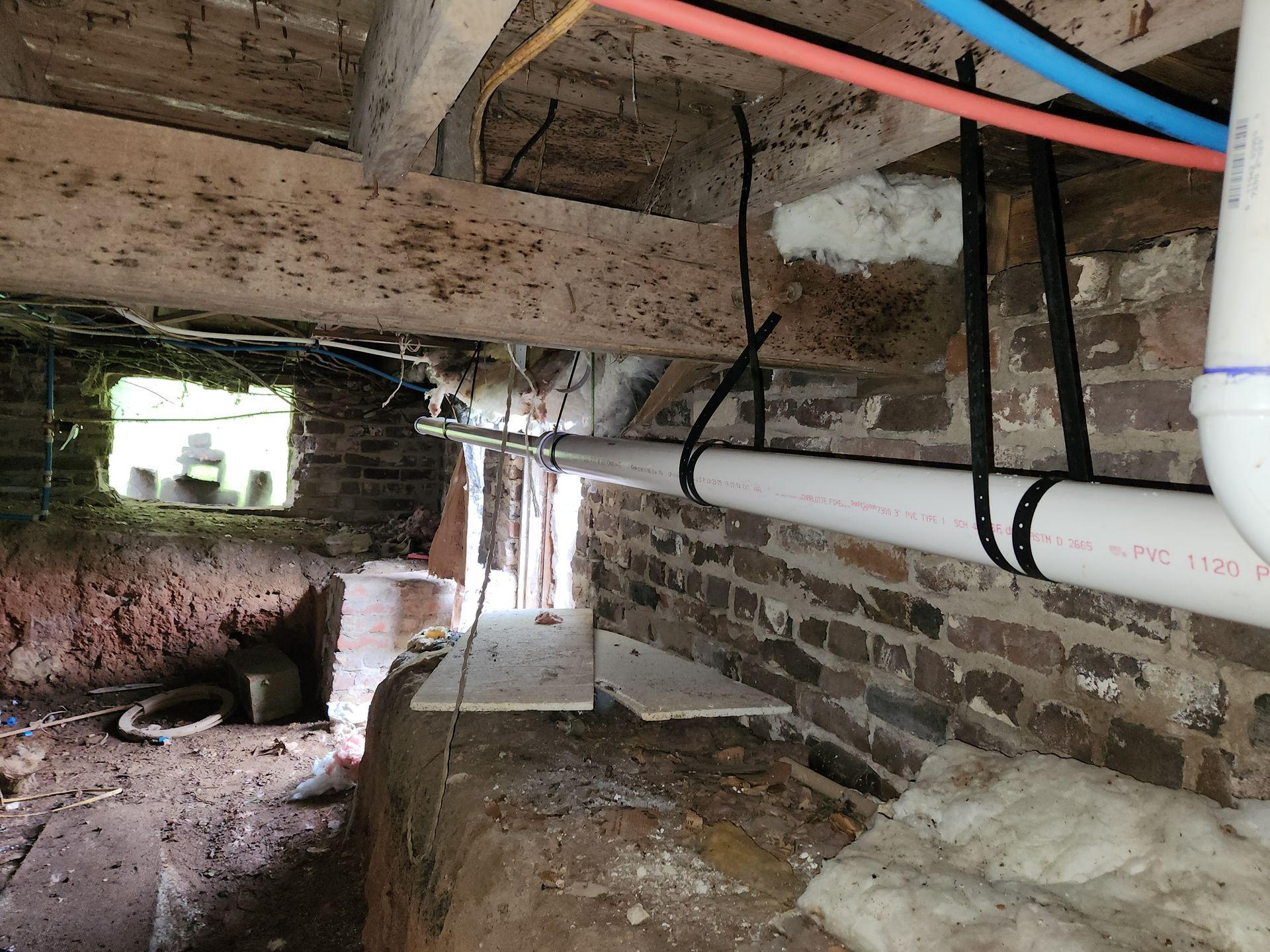 Crawlspace with exposed wooden beams, brick wall, and white PVC pipes under a house.