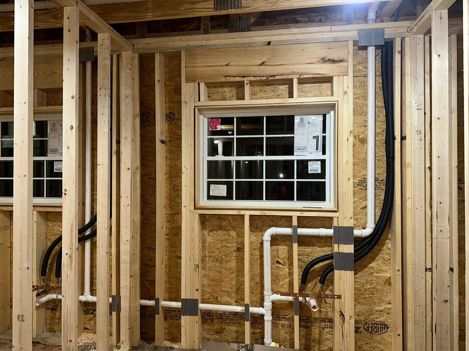 Interior wall framing with exposed wooden studs, pipes, and a small window during construction