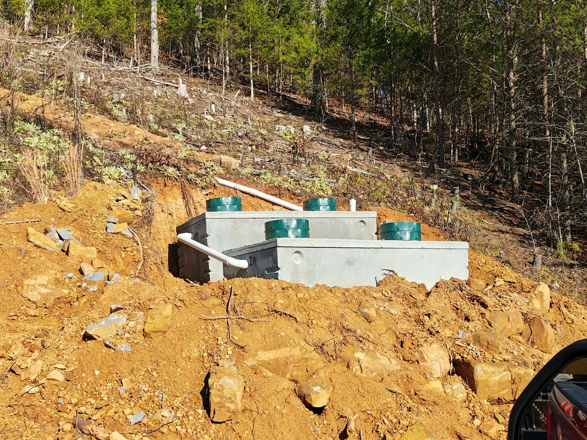 Concrete utility structure on a dirt hillside amid trees and rocks, with teal pipes visible.