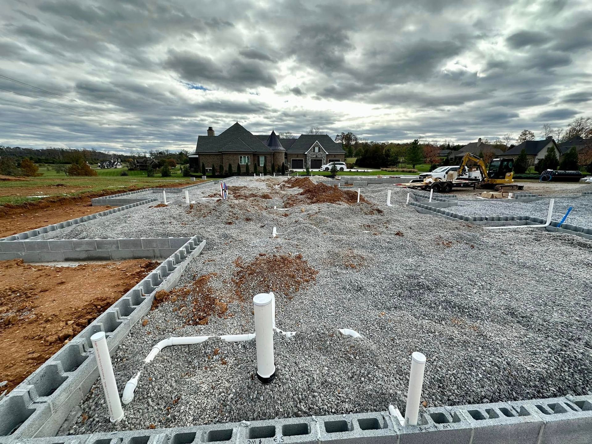 Gravel-covered construction site with foundations, pipes, and a large house under a cloudy sky
