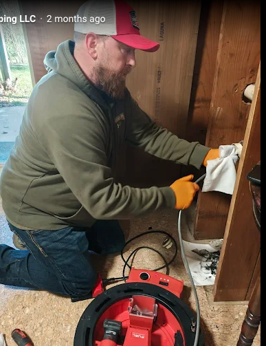 Worker in green hoodie and red cap kneeling, wiping inside a wooden cabinet beside a red vacuum tool.