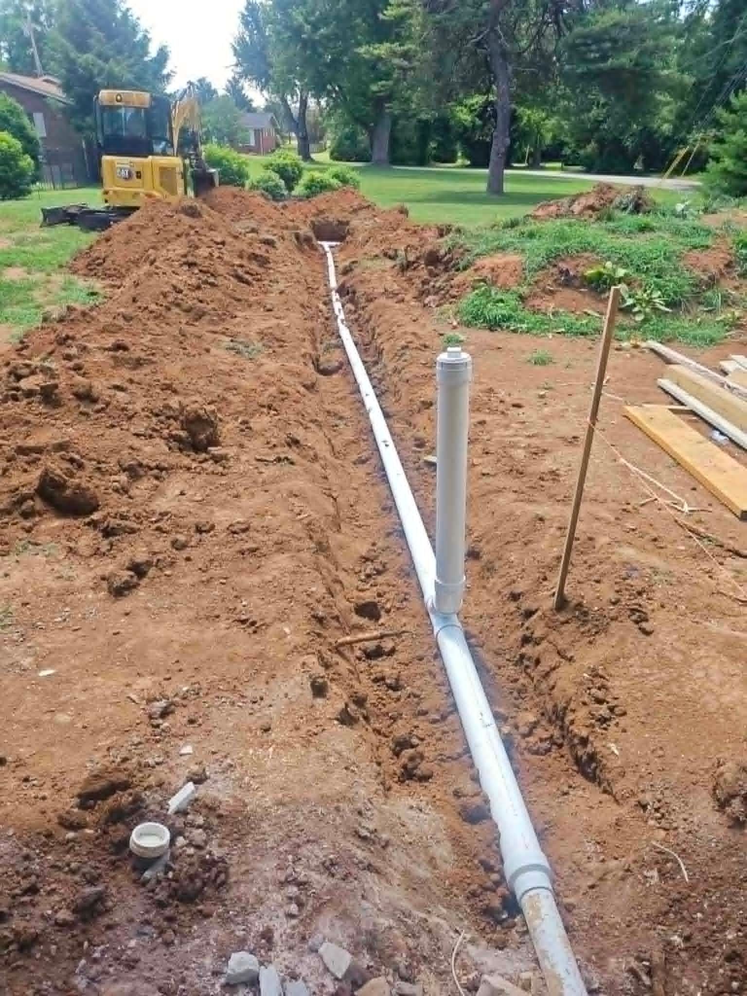 Trenched yard with white PVC pipe laid in dirt, construction equipment in background, and a small utility post.