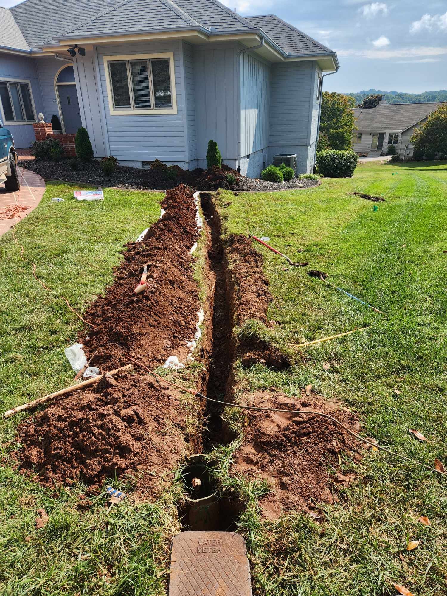 Trench dug across front yard beside a blue house, with soil piled along the cut line.