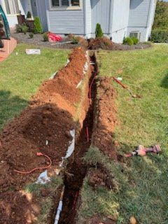 Large trench dug across a front lawn beside a house, exposing soil and a buried cable line.