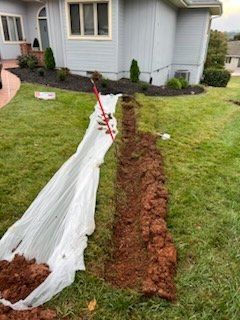 Plastic sheeting over a narrow trench beside a house, with a red marking flag and fresh soil along the yard.