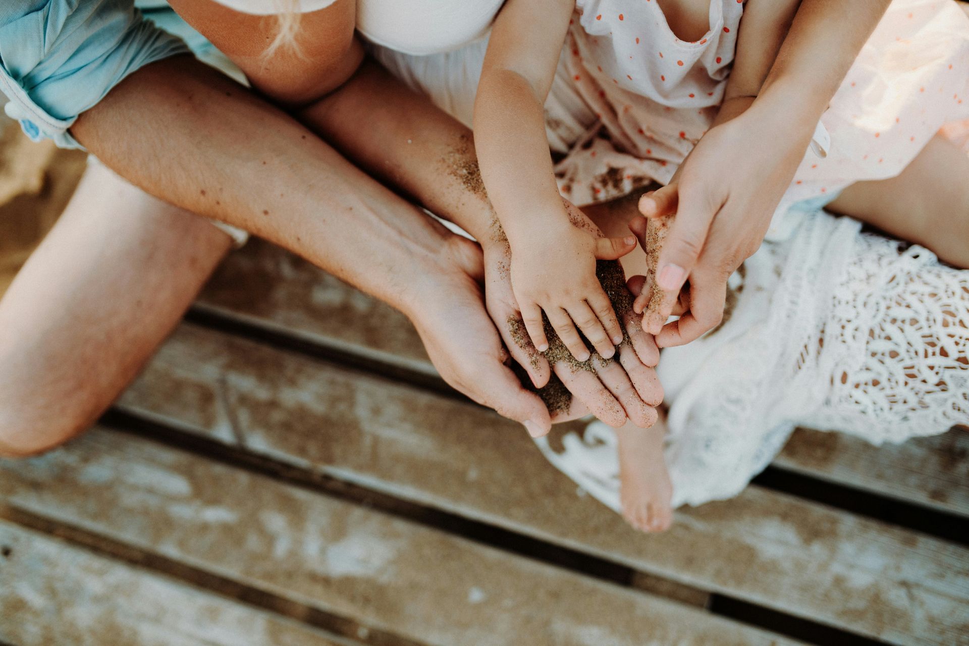 A family is sitting on a wooden bench with their hands in the sand.