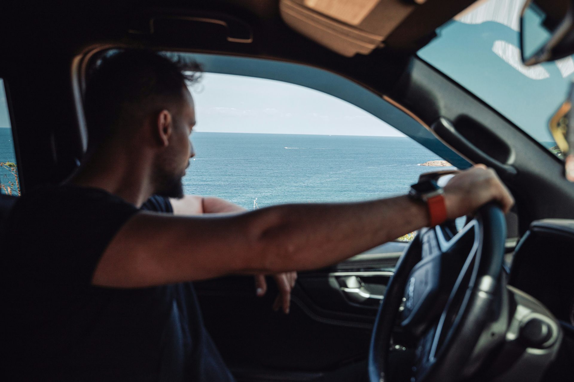 A man is sitting in a car looking out the window at the ocean.