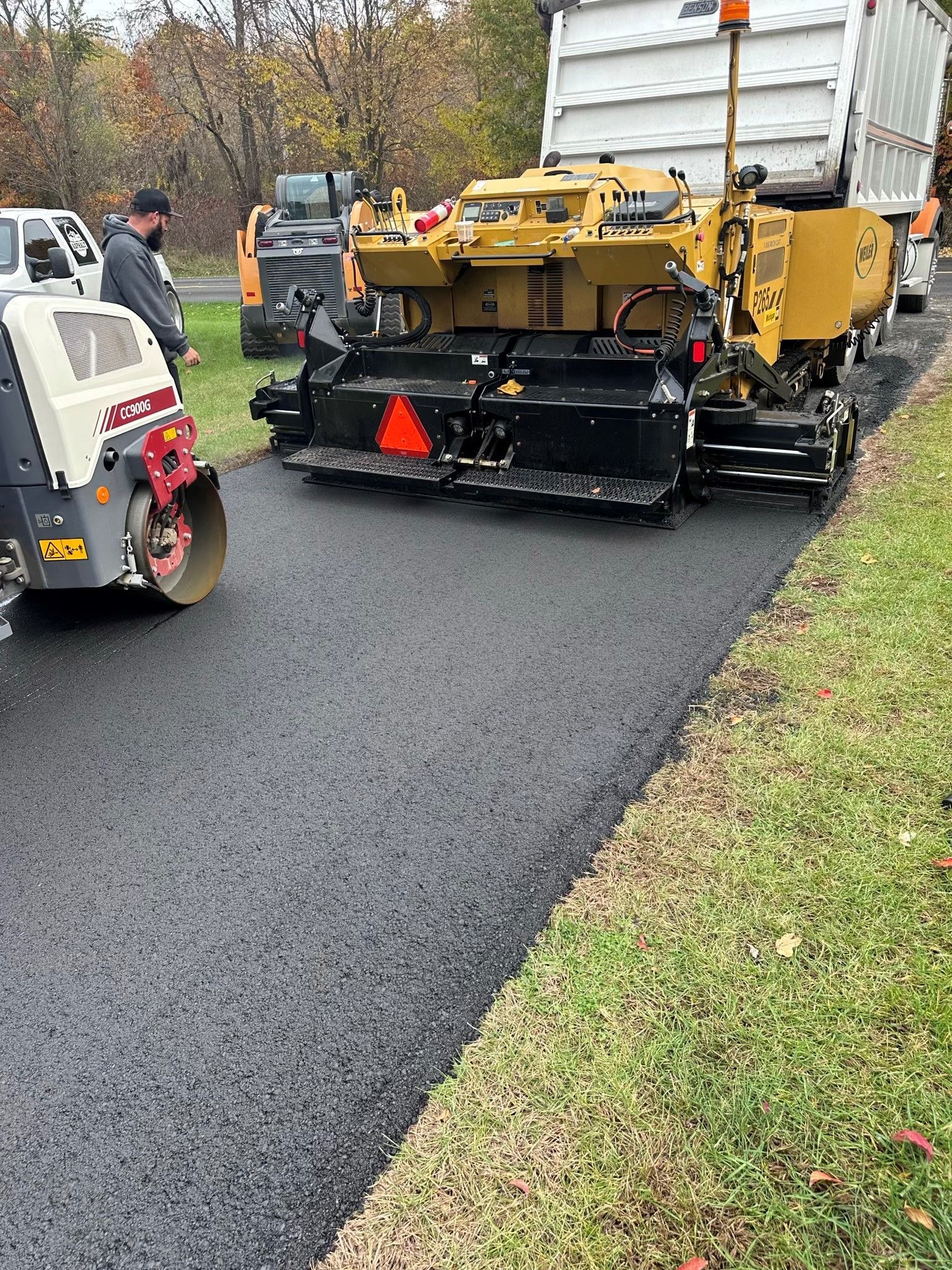 Road paver laying fresh black asphalt beside a white truck and workers on a roadside.