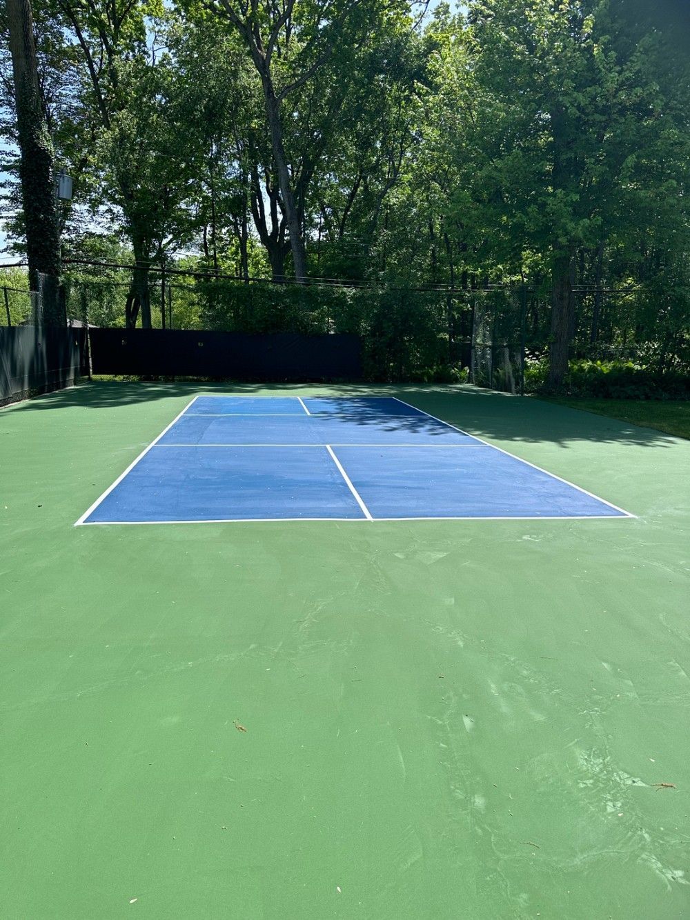 Outdoor tennis court with a blue playing surface and green surrounding area, bordered by trees.