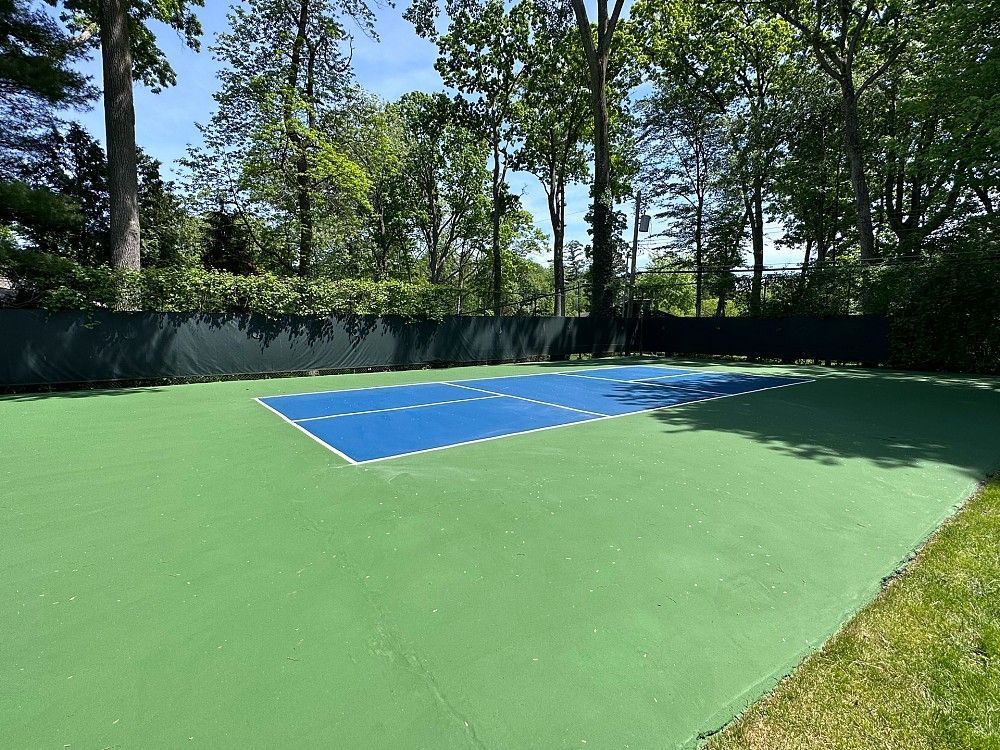 Outdoor tennis court with blue playing surface and green surrounding area, bordered by trees and fencing.