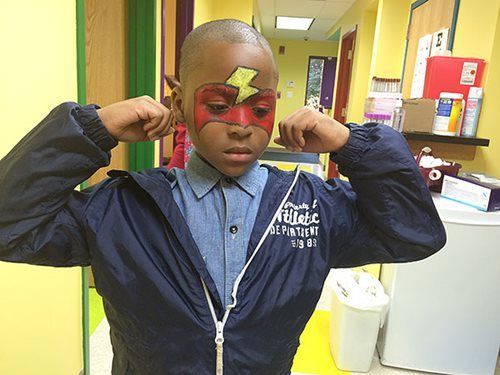 Child in a hallway wearing a superhero mask and jacket, flexing both arms.
