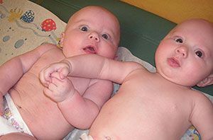 Two babies lying side by side on a blanket, holding hands and looking up with wide eyes
