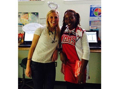 Two smiling students pose in a classroom, one in a red cheer uniform and one wearing a tiara.