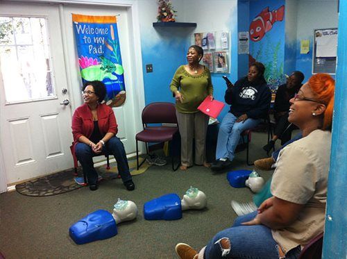 People gathered in a small room, seated and standing around blue plastic bowling pins and balls.