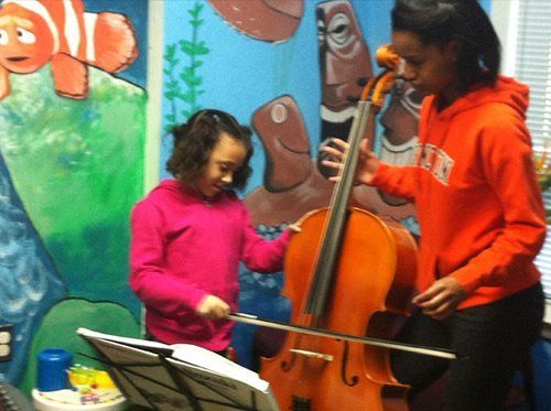 Two people playing a cello together in front of a colorful mural