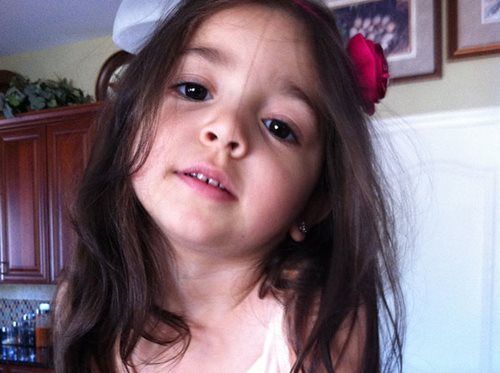 Close-up of a young child with long dark hair and red hair clips, leaning in indoors, looking at the camera