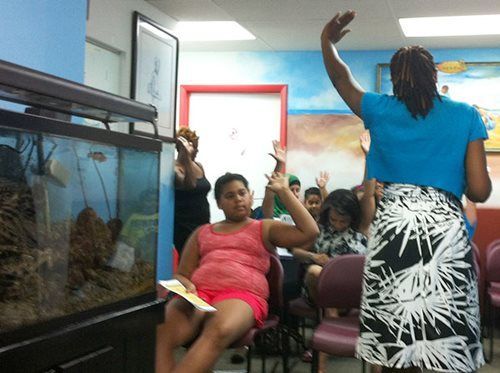 People seated indoors in a room, one woman standing and raising her hand near an aquarium.