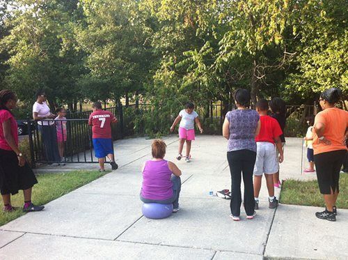 People exercising outdoors in a park, standing in a circle near trees and a fence.