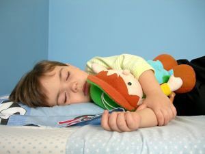 Child sleeping while hugging a plush toy on a bed, against a blue wall.