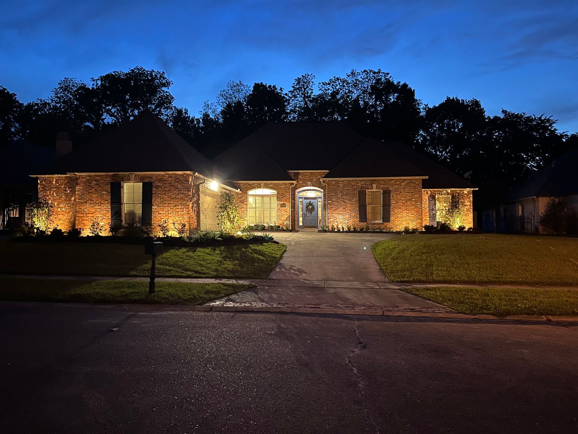 A brick house at dusk is illuminated by warm-toned lights. A dark sky forms a backdrop.