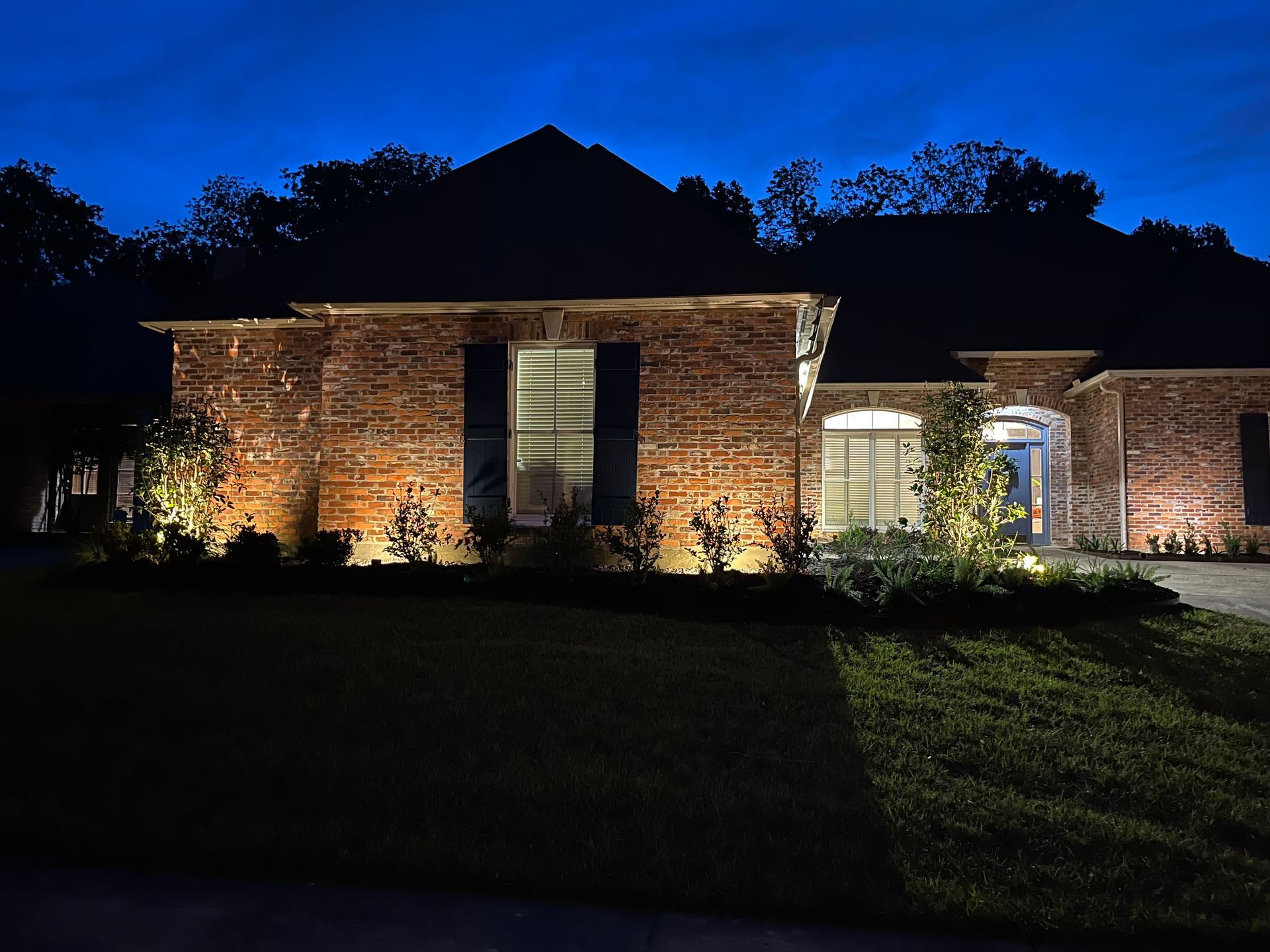 Brick house illuminated at night with landscape lighting. Dark blue sky, green lawn, and shadowy trees.
