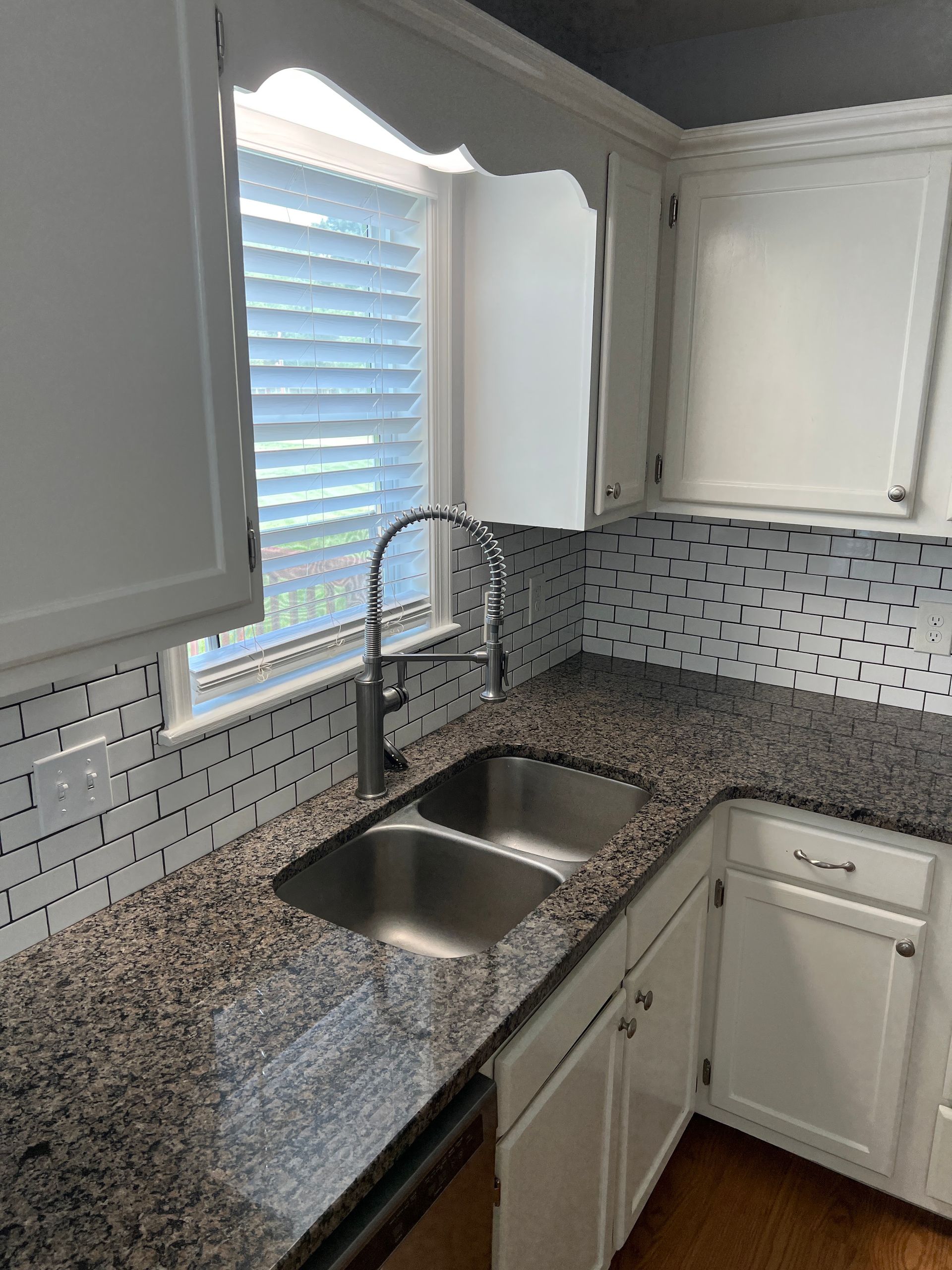Kitchen corner with white cabinets, granite counters, tiled backsplash, and stainless double sink by a window