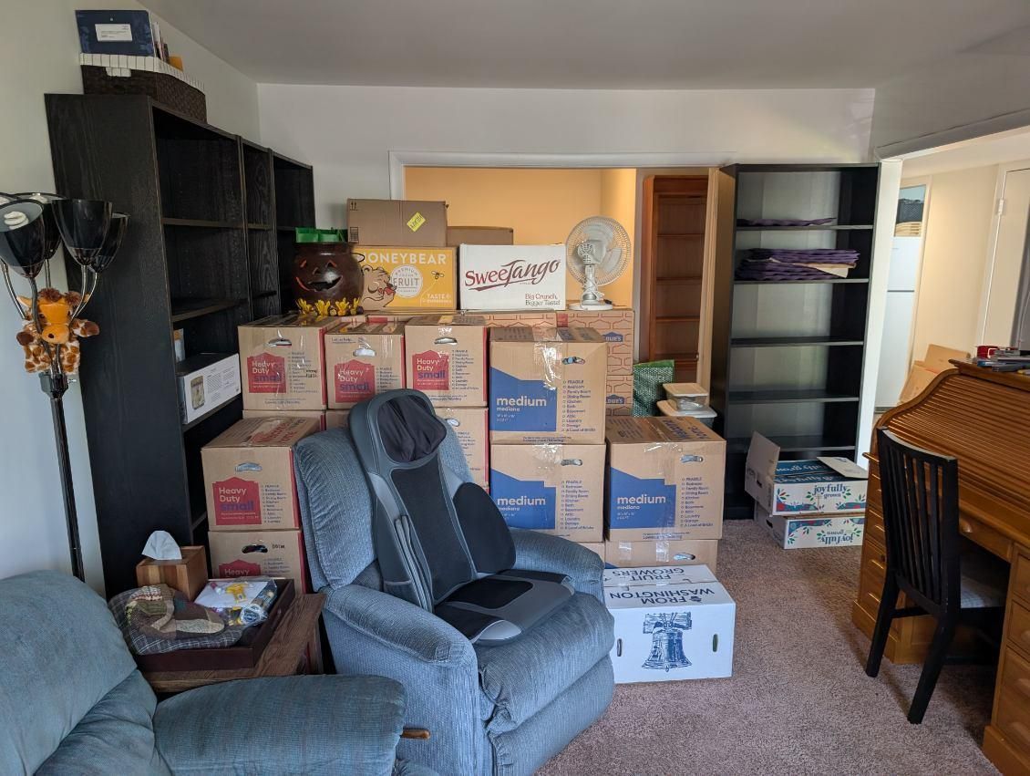 Cluttered living room with stacked moving boxes, a gray recliner, shelving, and a desk near a window.